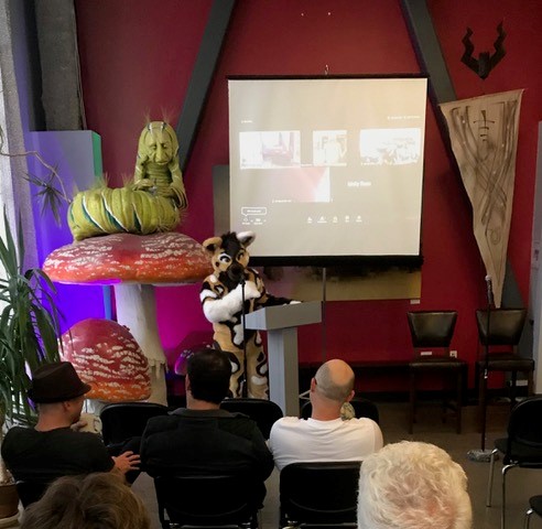 Presentation occuring with presenter dressed up as a Furry, and a large caterpillar sitting on a mushroom behind them. Audience sitting in chairs with backs to camera.