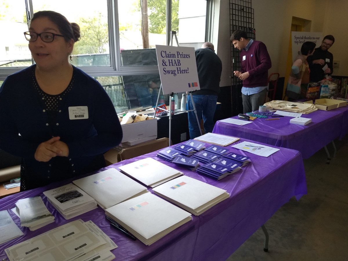 Person with hair in bun with glasses standing behind table with purple table cloth with name tags, folders, booklets, and next to sign that says "Claim Prizes & HAB Swag Here!" Adjacent table with purple table cloth and 4 people standing behind it on right.