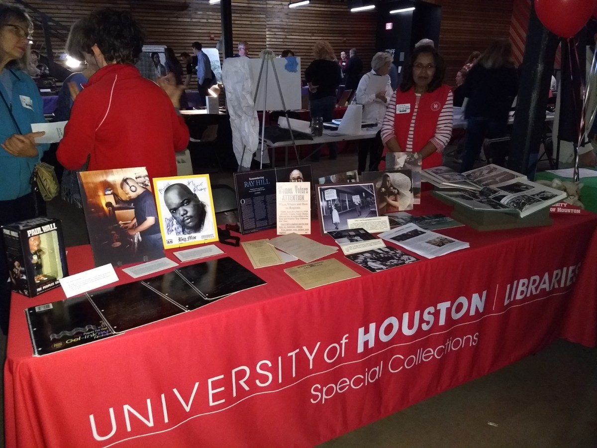 Table with red table cloth that says "University of Houston Libraries Special Collections" on it with people standing behind table and different textual and photographic documents displayed on table.