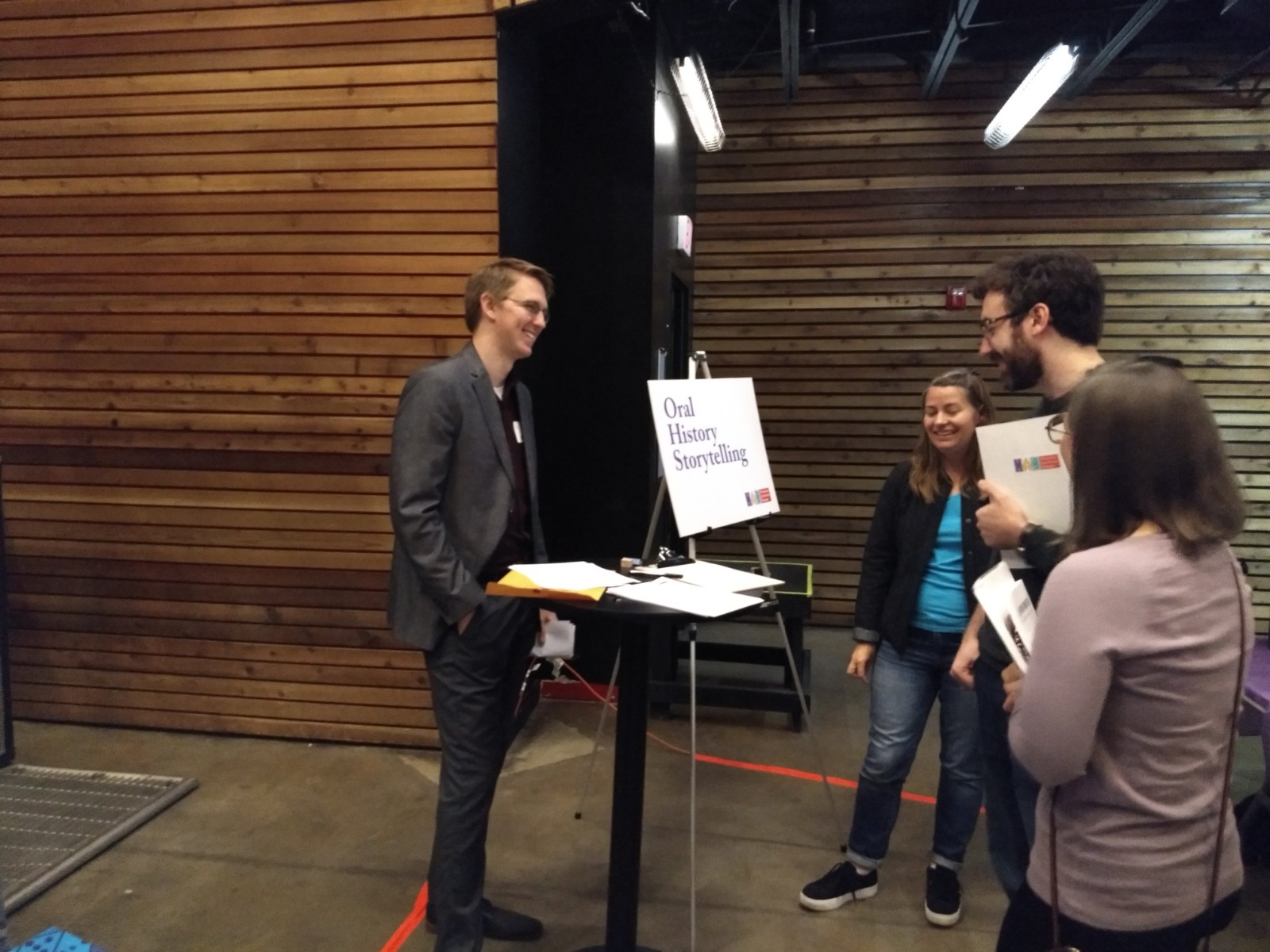 People standing around sign that says "Oral History Storytelling" and table with clipboards on it.