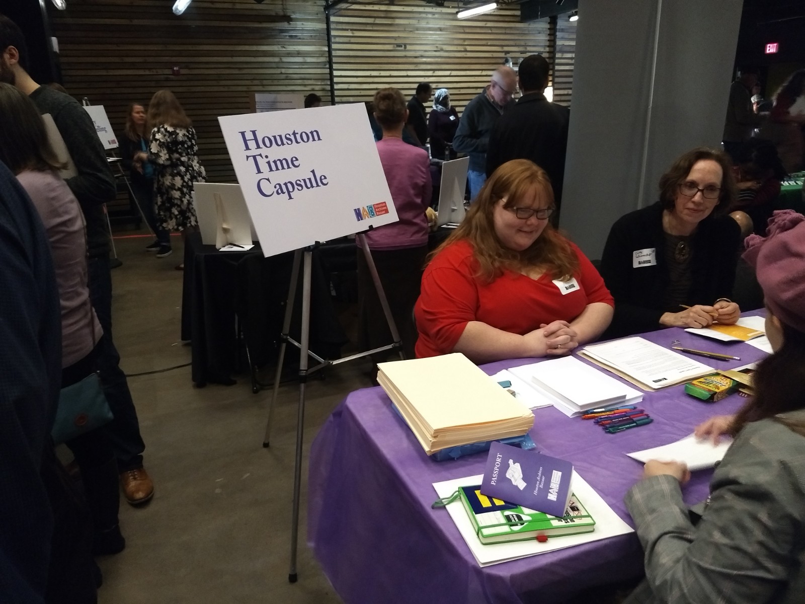 Two people sitting behind table with purple table cloth with standing sign that says "Houston Time Capsule" standing to the left, person in front of table sitting and engaging.