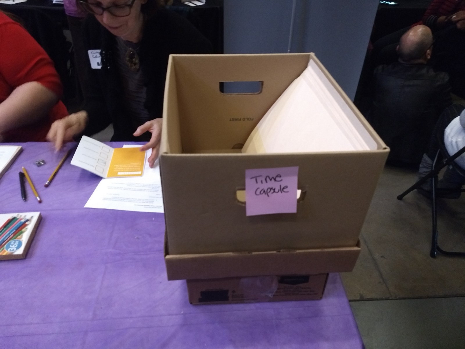 Close up of brown box with sticky note with "Time Capsule" written on it, sitting on table with purple table cloth.